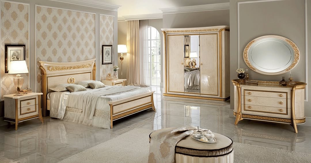 Elegant Italian bedroom with ornate cream and gold furniture, including a bed, nightstands, wardrobe, dresser with round mirror, and upholstered bench on marble tile flooring.
