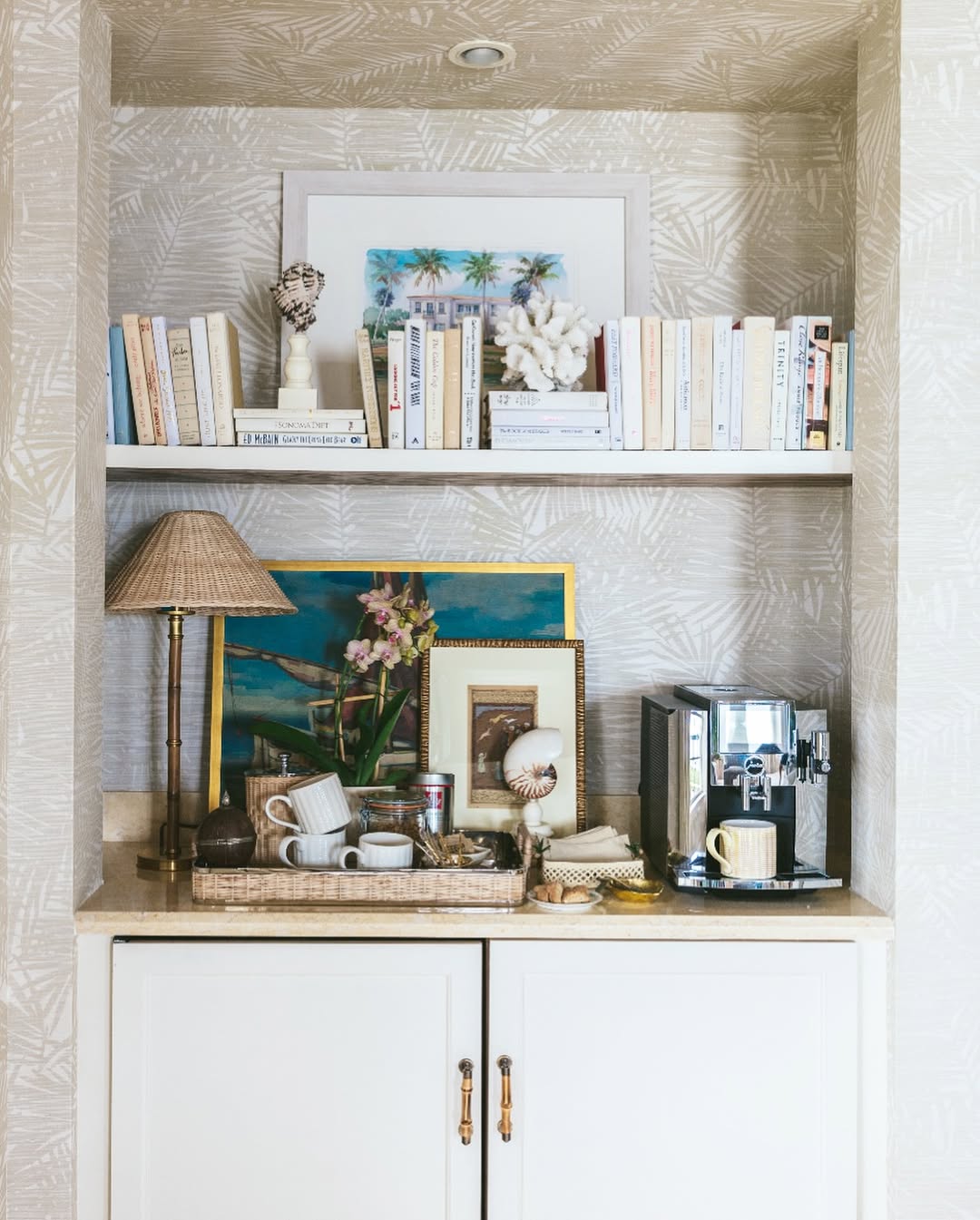 A cozy coffee bar nook with books, framed art, decorative objects, a coffee machine, cups, and a lamp above white cabinets—perfect for stylish Kitchen coffee bar ideas that help you escape countertop chaos.