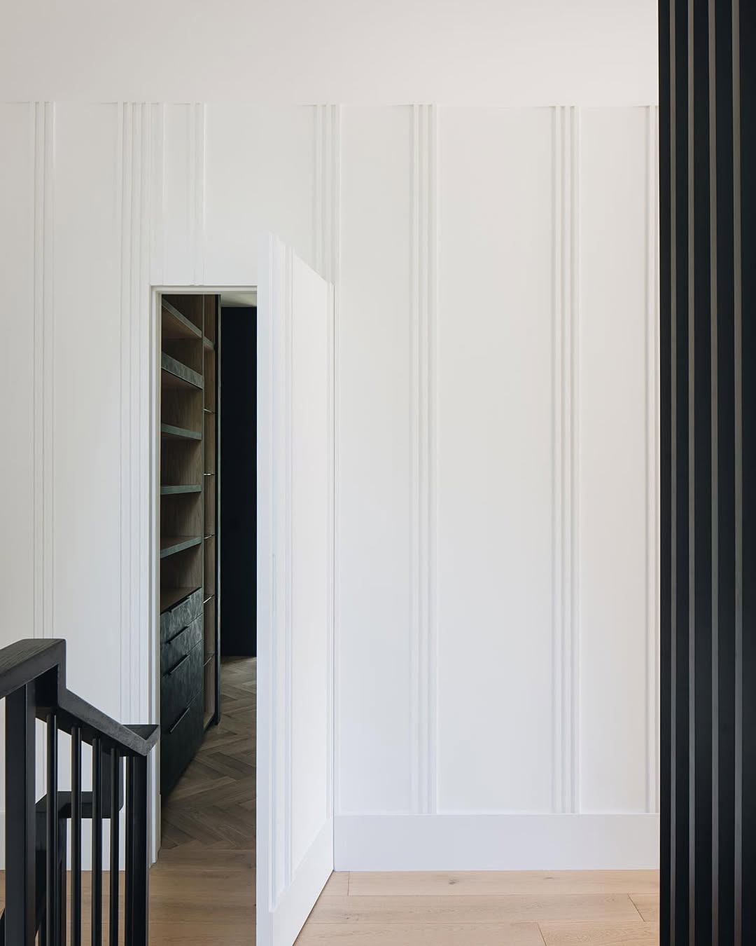 A minimalist interior features white paneled walls, a partially open hidden door leading to shelving, light wood flooring, and a black railing in the foreground—perfect inspiration for stylish homeowners seeking subtle hidden door ideas.