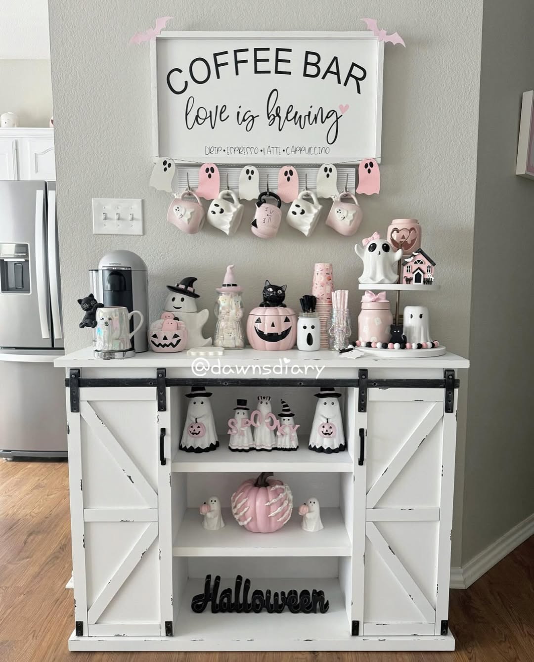 A white coffee bar with a Halloween theme, decorated with pink and white ghost and pumpkin ornaments, mugs, and a sign that reads “COFFEE BAR love is brewing”—perfect inspiration for kitchen coffee bar ideas without any countertop chaos.