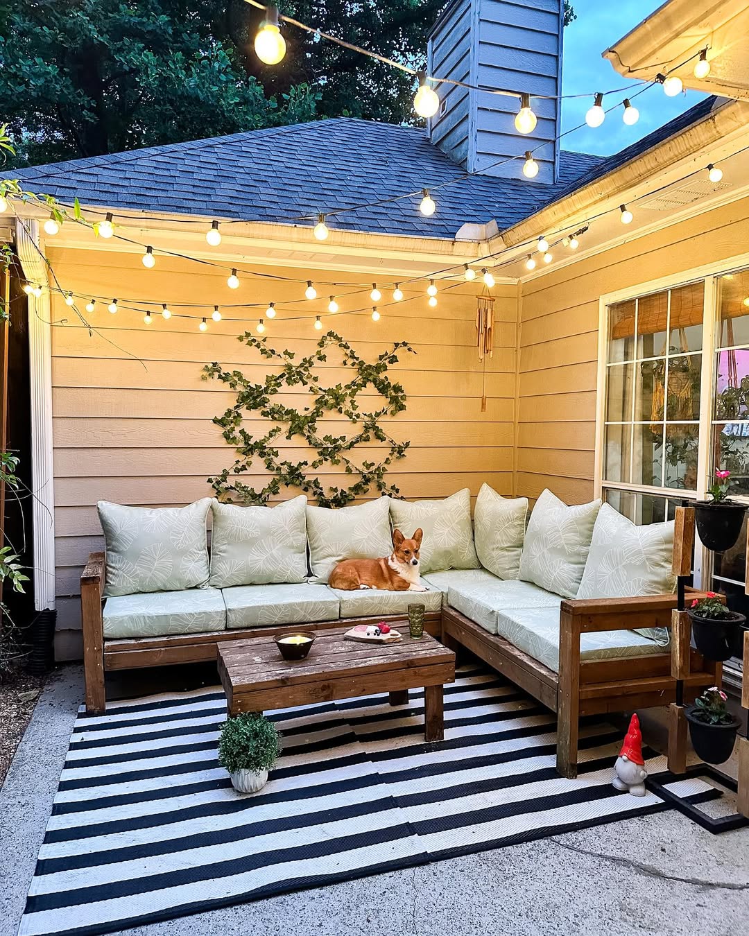A corgi lies on a light green outdoor sectional under string lights on a patio, surrounded by stylish patio decoration like a striped rug, wooden table, and potted plants.