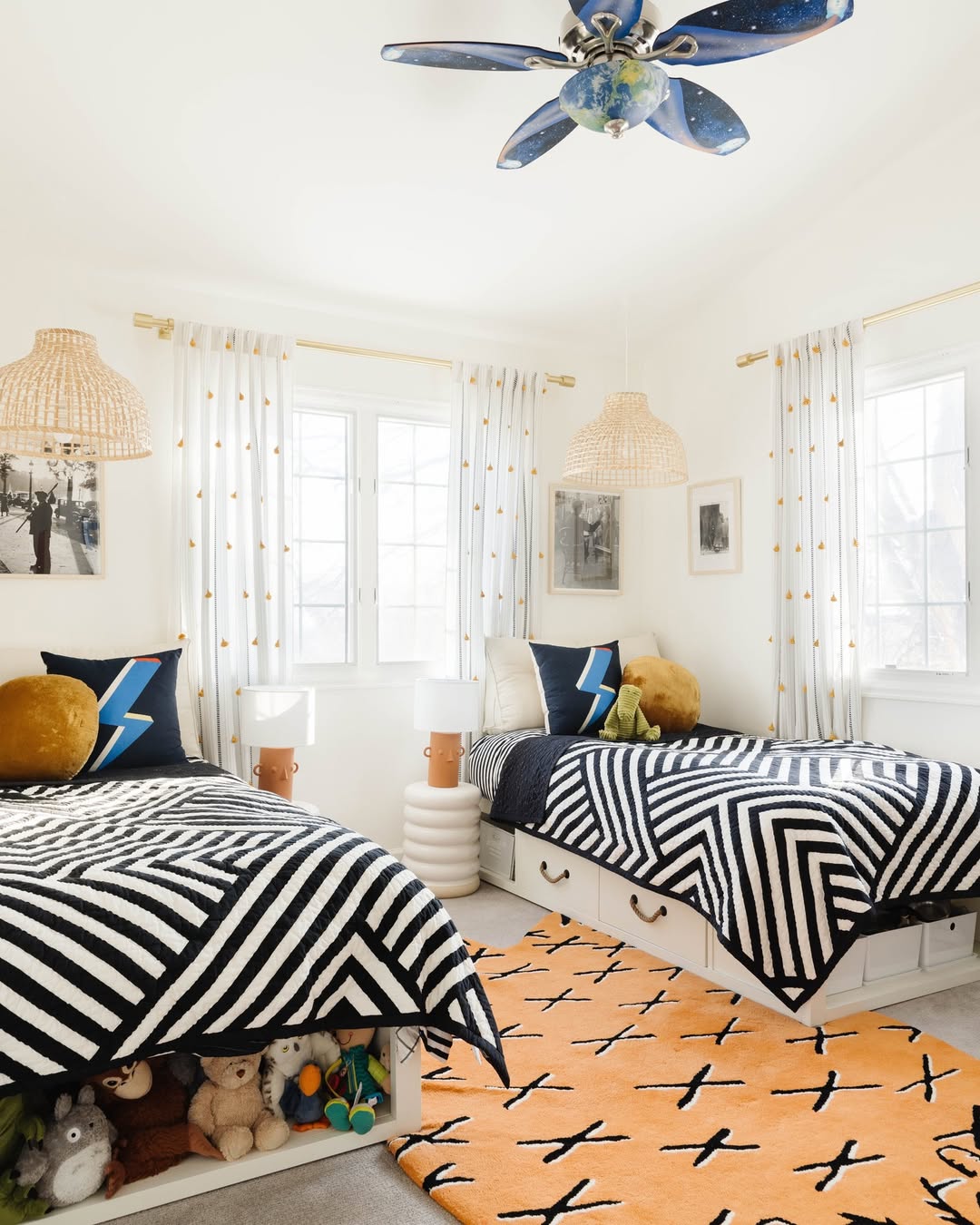 A bright boys’ bedroom idea featuring two twin beds with geometric black-and-white bedding, cozy stuffed animals, clever storage, an orange patterned rug, and a ceiling fan with a globe design—perfect for sleep and play.