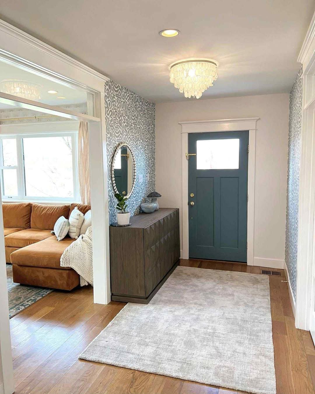 A bright modern entryway with a blue front door, wooden sideboard, round mirror, patterned accent wall, chandelier, and a light area rug on hardwood floors—perfect for fresh entryway ideas.