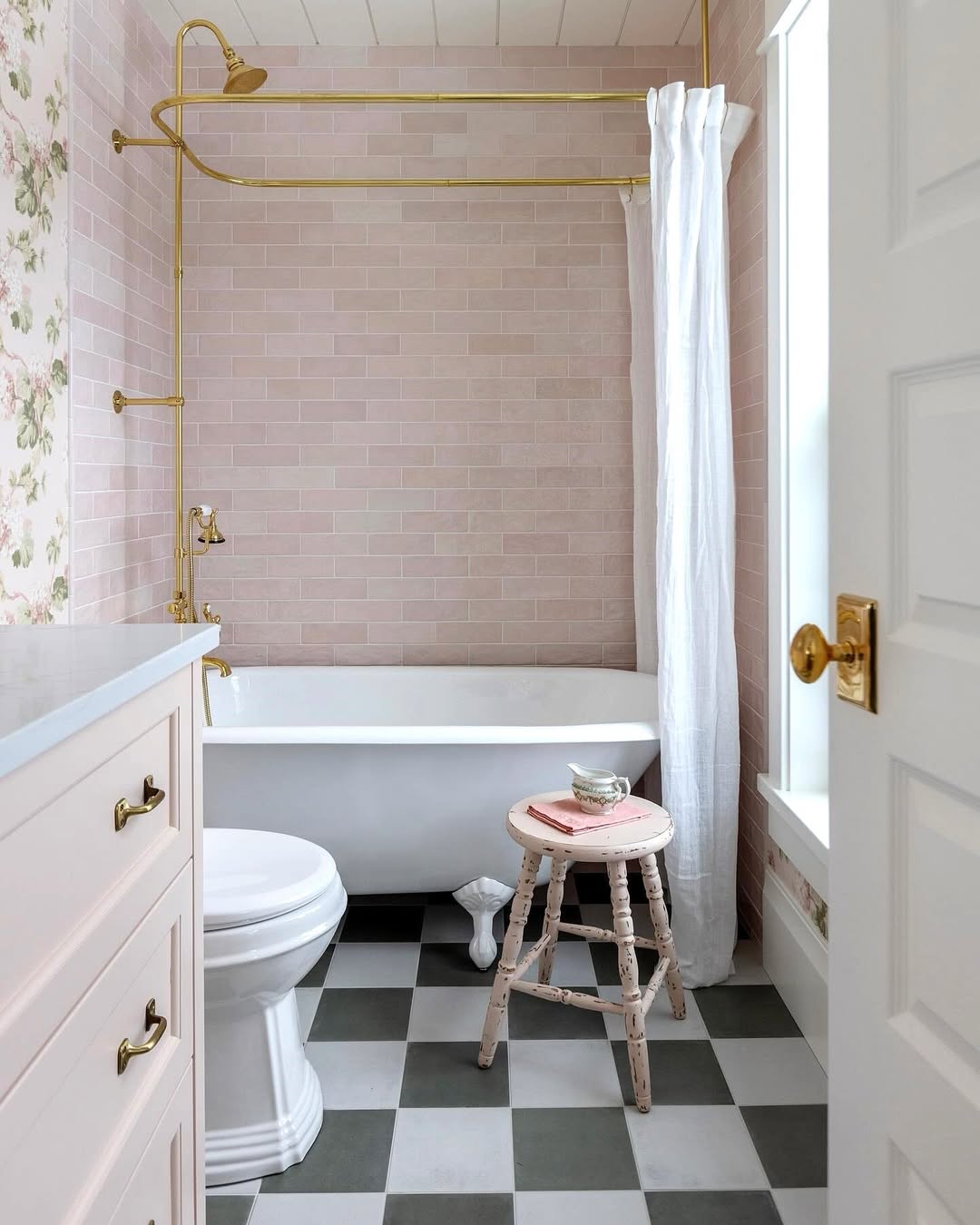 A bathroom with pink tile walls, a clawfoot bathtub with gold fixtures, a white shower curtain, checkered black-and-white floor, and a wooden stool holding a small tray and cup—perfect inspiration for charming Bathroom Tub Ideas.