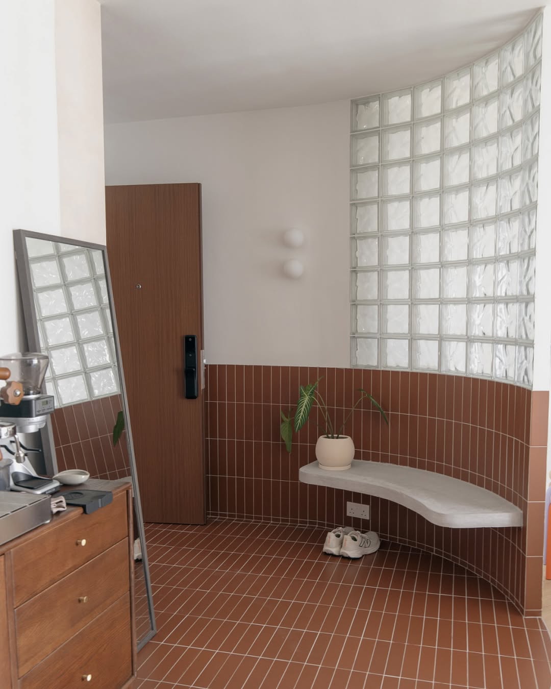 Modern Entryway Ideas come to life in this space, featuring brown tile flooring and walls, a curved glass block partition, floating bench, potted plant, mirror, and a small cabinet with coffee machine for practical entryway decor.