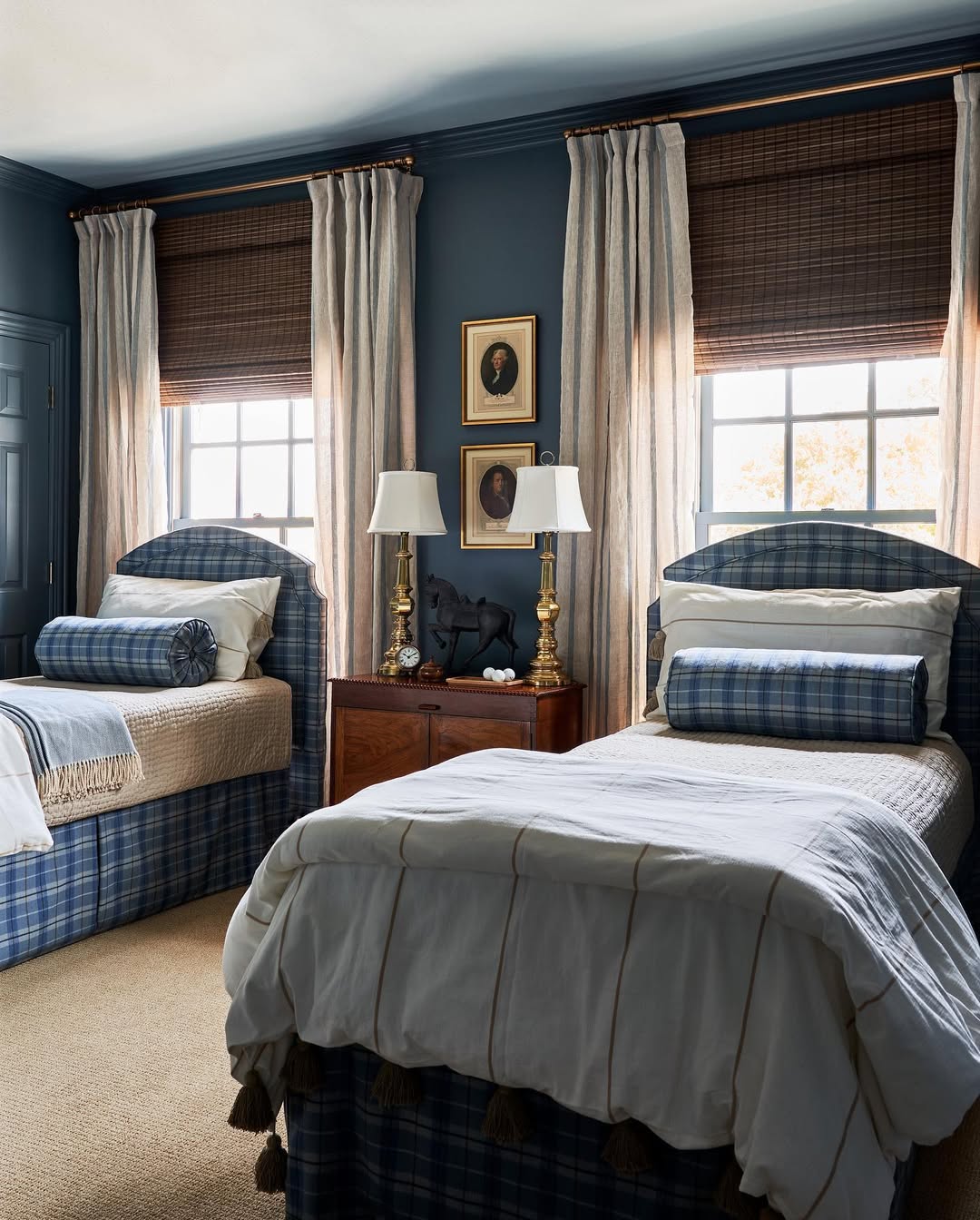 Two twin beds with blue plaid upholstery and white bedding are placed side by side in a blue-walled bedroom, featuring clever storage solutions—a smart pick for boys bedroom ideas—with two windows, drapes, a wooden nightstand, lamps, and framed art above.
