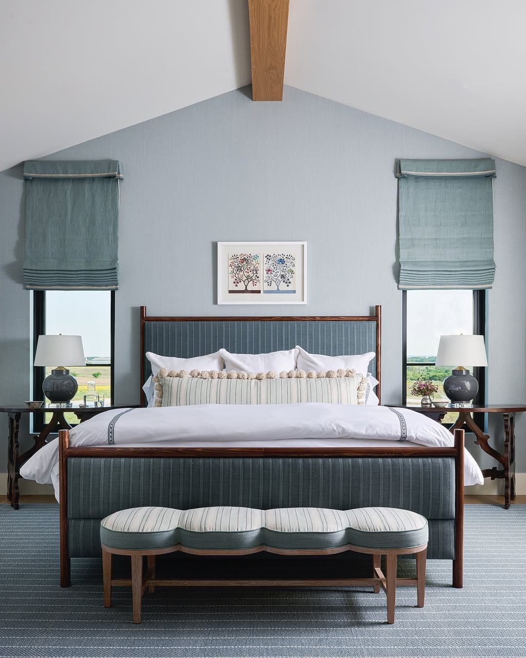 A modern bedroom with a blue upholstered bed, white bedding, two bedside tables with lamps, a striped bench, and two windows with blue Roman shades. Artwork hangs above the bed, offering fresh inspiration for ranch house decor.
