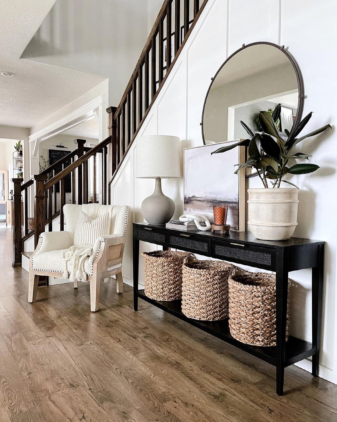 A hallway with a staircase, a console table with baskets, a lamp, a plant, and a round mirror creates inviting hallway decor alongside a white armchair with a blanket and pillow.