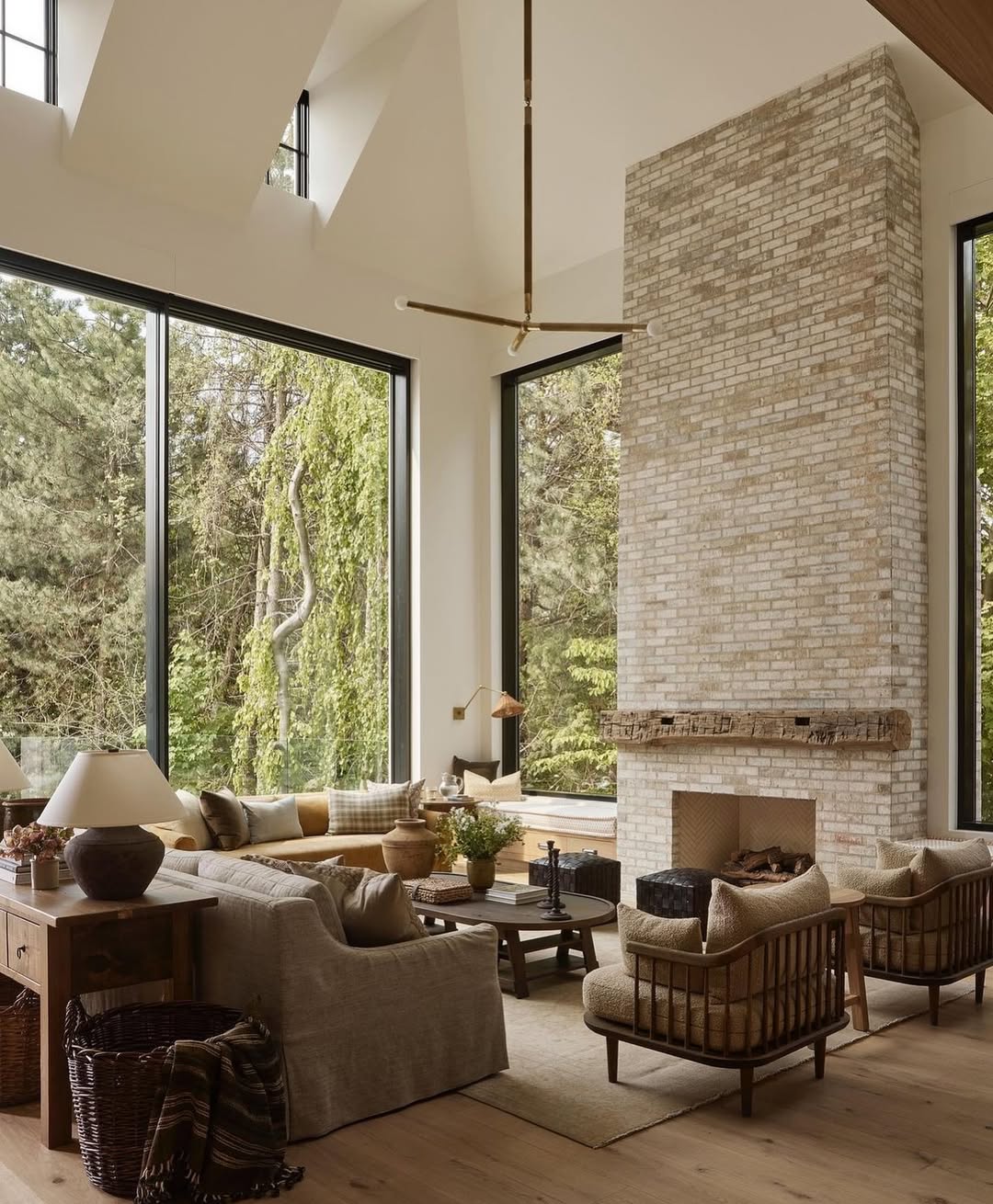 Modern high ceiling living room with large windows, neutral-toned furniture, a tall white brick fireplace, wooden accents, and views of trees outside—perfect inspiration for your living room ideas.
