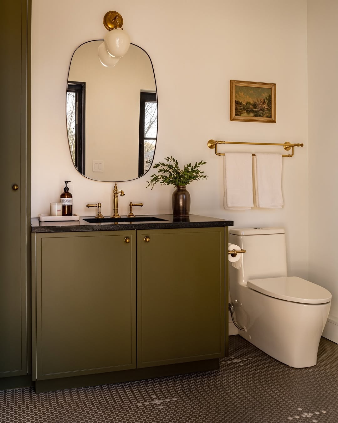 This stylish powder room features an olive green vanity, black countertop, oval mirror, wall sconce, toilet, towel rack with two white towels, and small framed artwork on cream-colored walls—perfect for those seeking wow-factor powder room ideas.