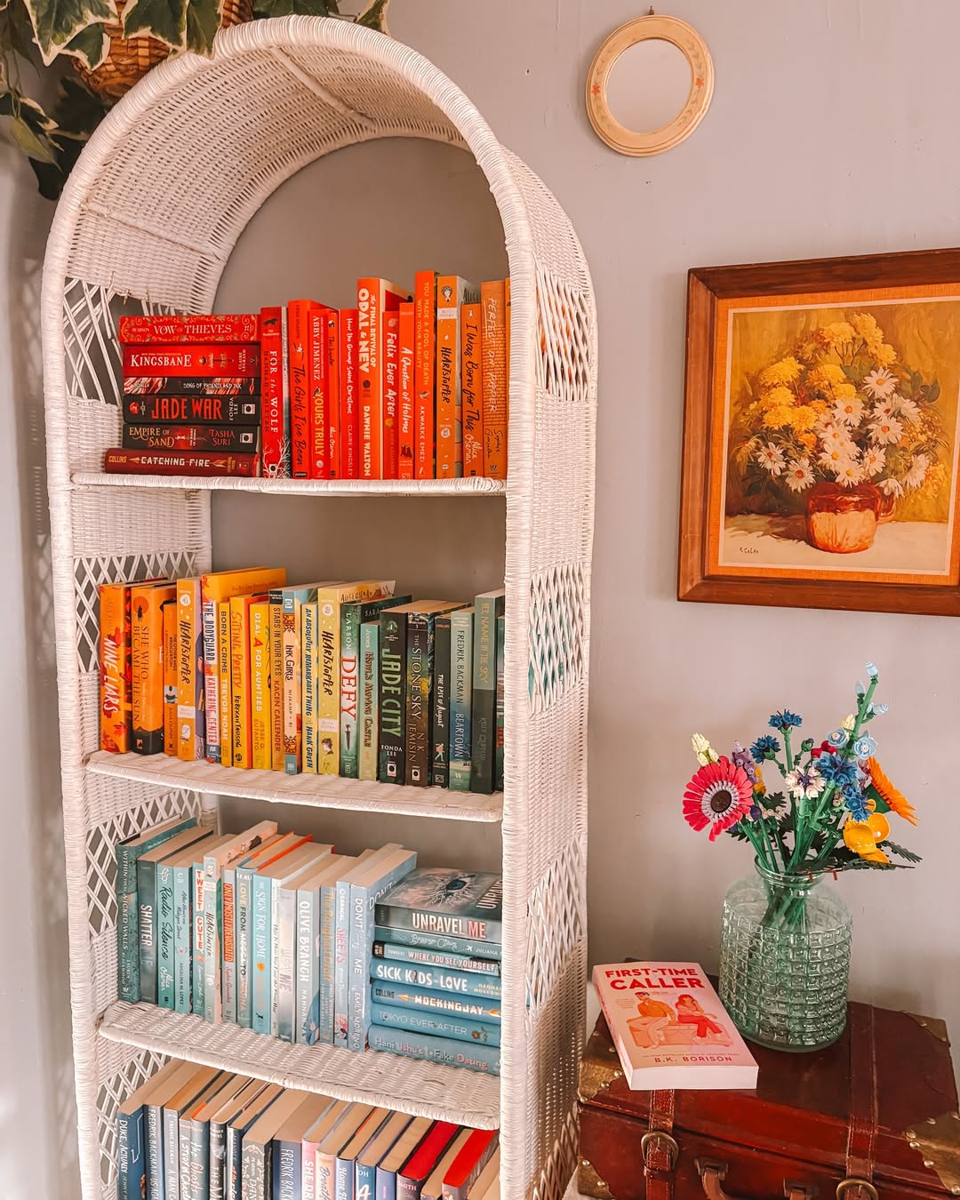 A white wicker bookshelf holds books arranged by color, from red to blue, showcasing creative bookshelf styling with a vase of flowers, a painting, and a book on a nearby table.