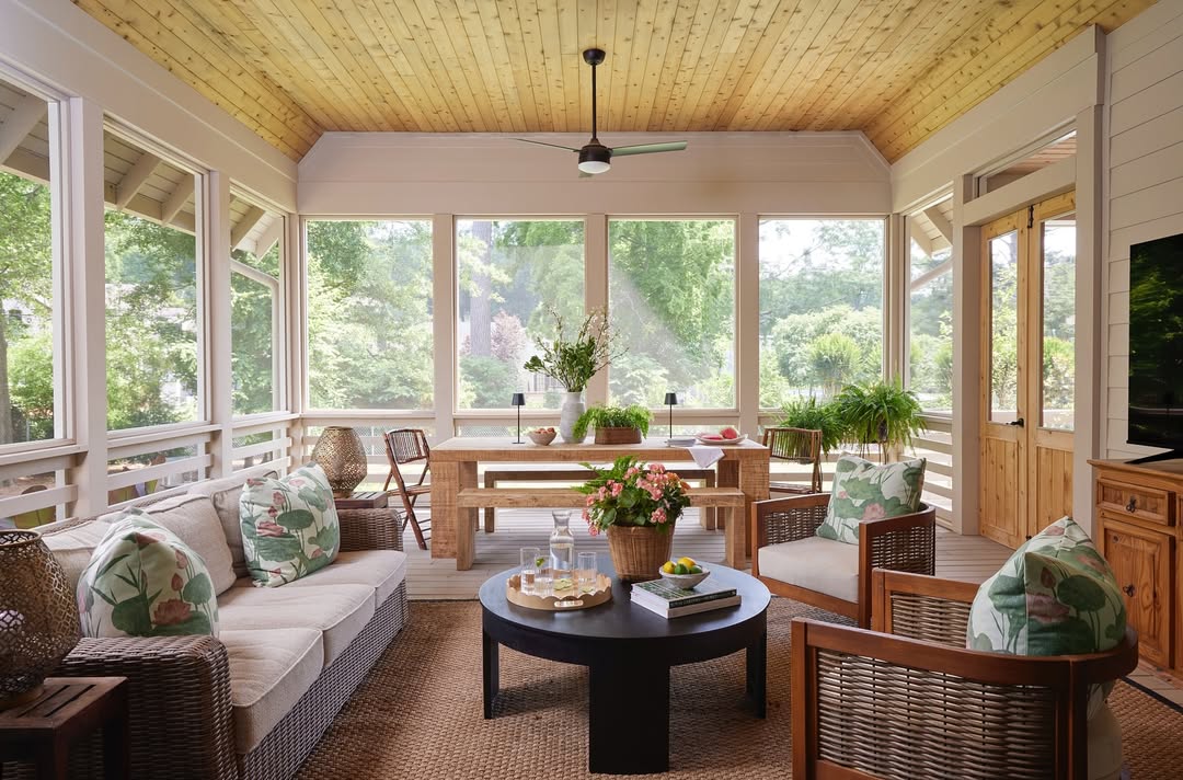 A bright, screened-in seasonal porch features wicker sofas with floral cushions, a round coffee table, a wooden dining table, green plants, and large windows overlooking a lush garden.