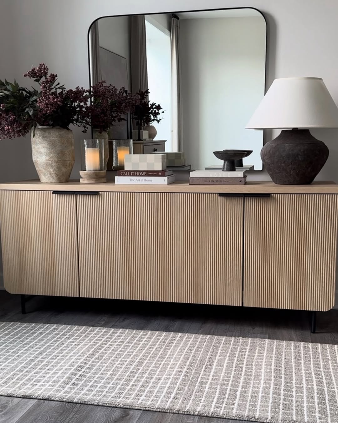 A wooden sideboard with books, candles, vases, and a lamp sits against a wall with a large rectangular mirror above it; a patterned rug lies on the floor, offering chic sideboard decor ideas for any space.