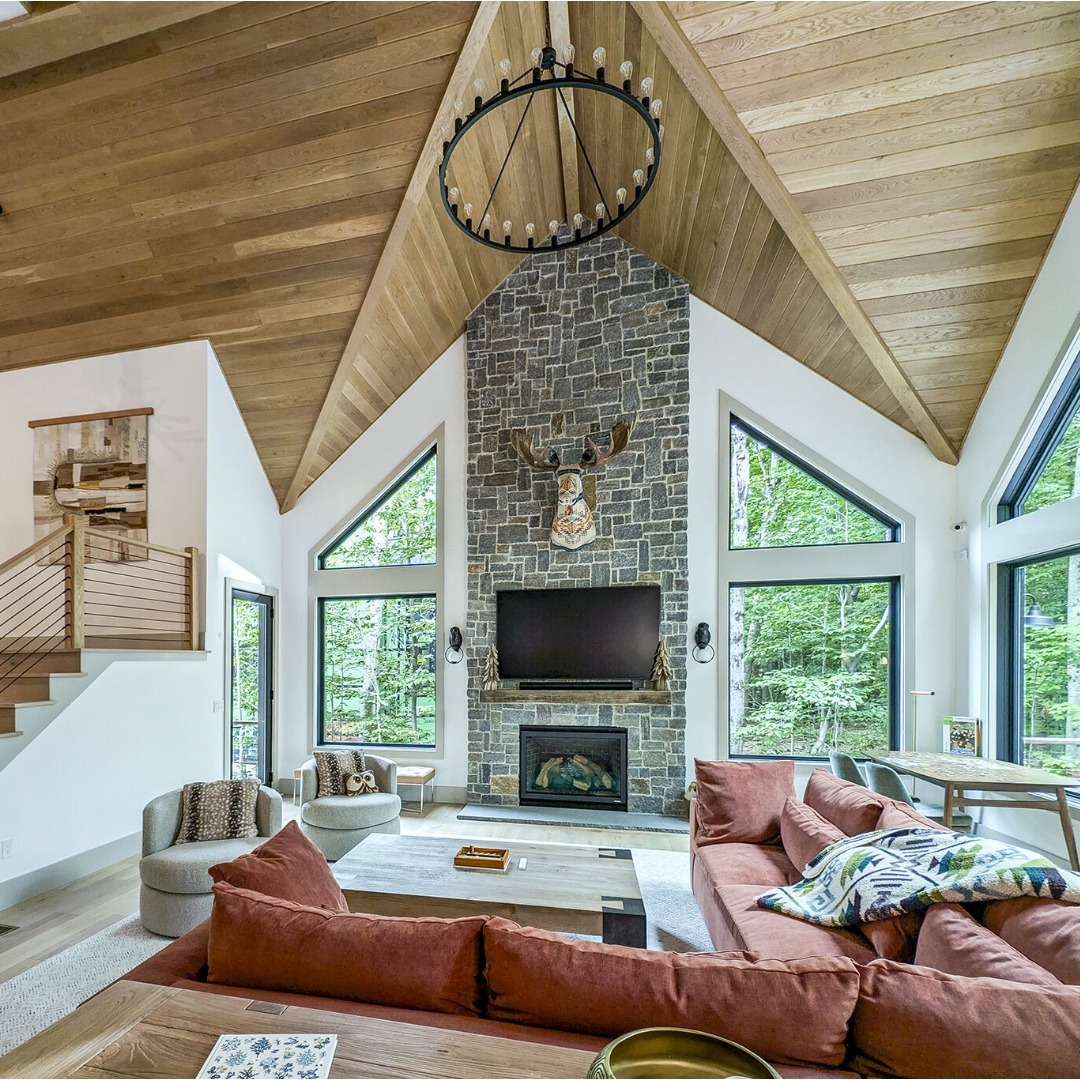 Modern high ceiling living room with vaulted wood ceiling, stone fireplace, mounted TV, and large windows. Red sectional sofa faces the fireplace; staircase is visible to the left—perfect inspiration for bigger home designs.