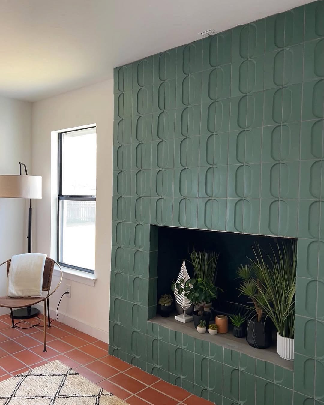 A modern living room with a green fireplace tile design, potted plants in the hearth, a window, a floor lamp, and a chair with a cushion.