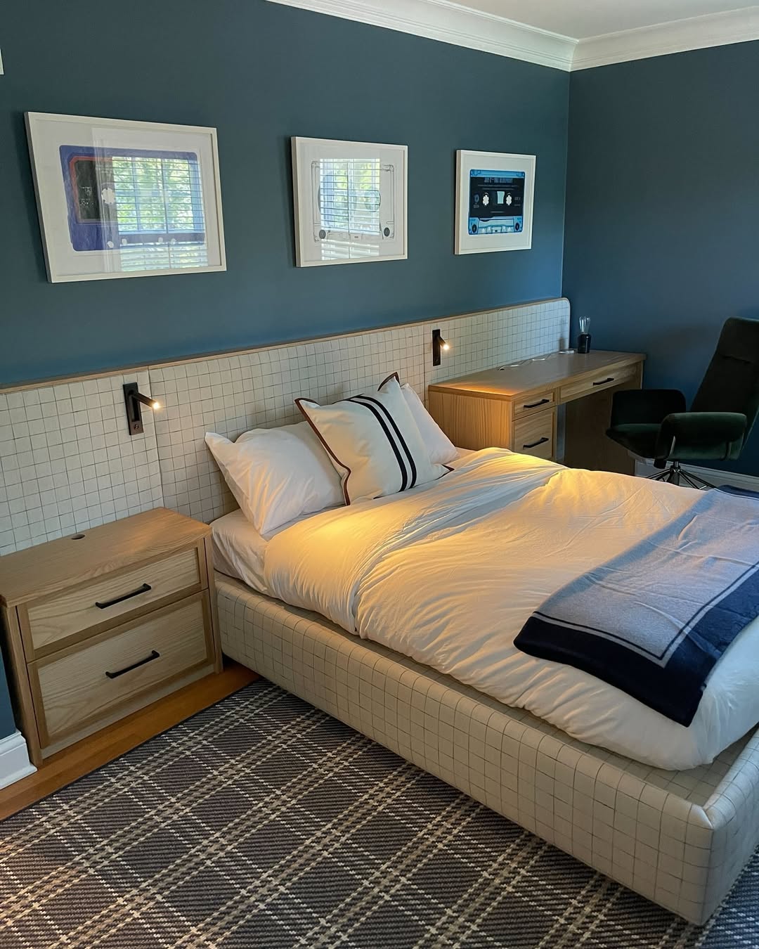A neatly made bed with white bedding and a blue blanket in a modern boys bedroom with blue walls, wooden furniture featuring smart storage solutions, wall-mounted lights, and framed artwork above the headboard.