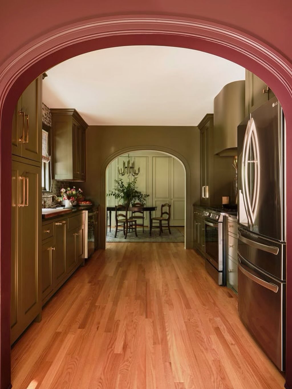 A kitchen with olive green cabinets, stainless steel appliances, and wood flooring showcases smart small kitchen design, featuring an arched doorway that leads to a dining room with a table and chairs.