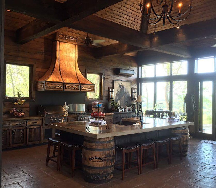 Rustic kitchen with wood walls, large island supported by barrels, metal range hood, bar stools, and large windows letting in natural light—perfect for those seeking rustic kitchen ideas.