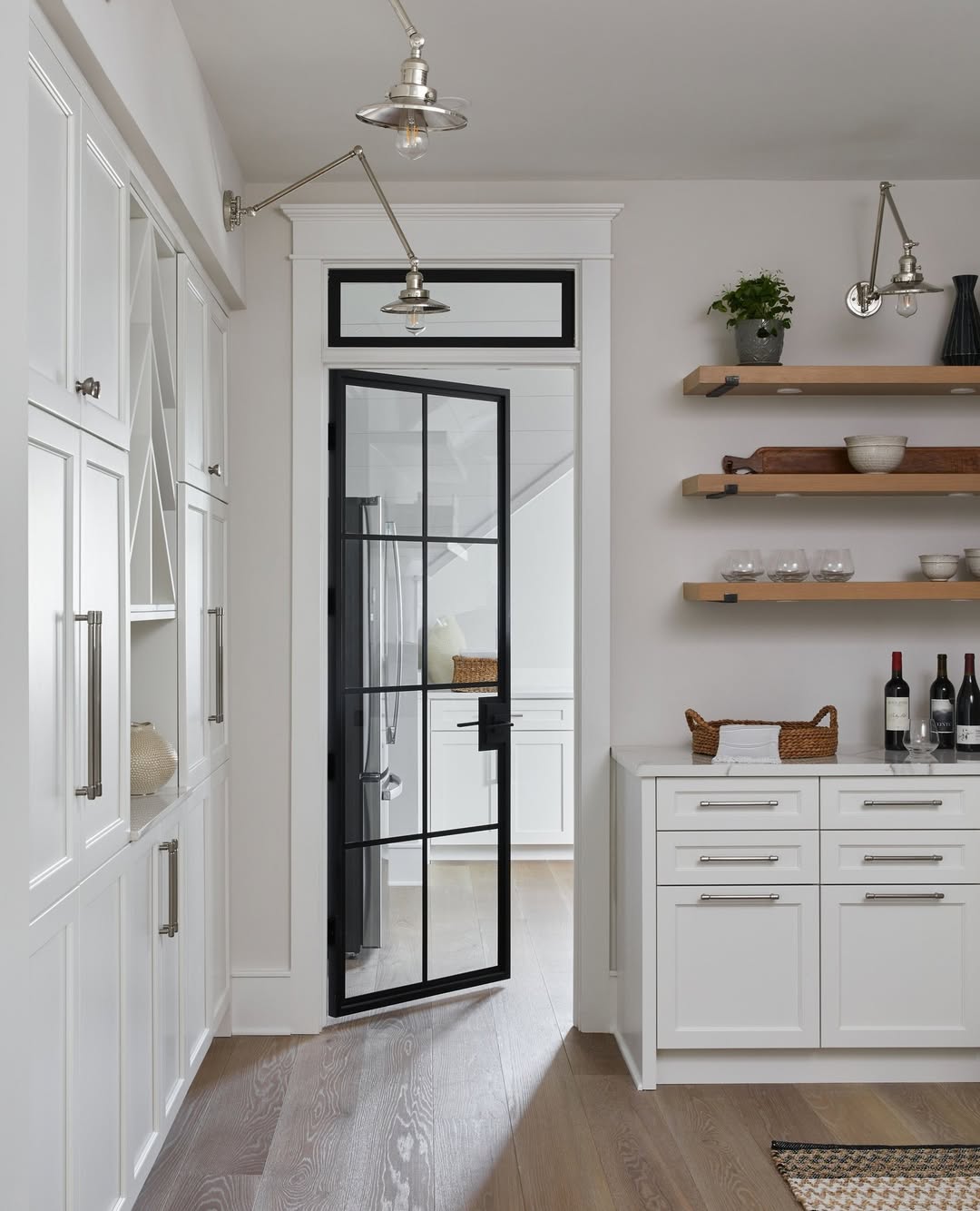 Modern kitchen with white cabinets, open wooden shelves, bottles on the counter, and a stylish black-framed glass pantry door leading to another room. Light wood flooring and metal lamps overhead. Perfect inspiration for pantry door ideas.