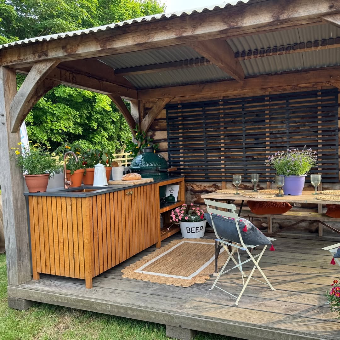 A charming rustic outdoor kitchen and dining area features wooden cabinets, potted plants, a table set for two, and a corrugated metal roof—perfect inspiration for those seeking outdoor kitchen ideas.