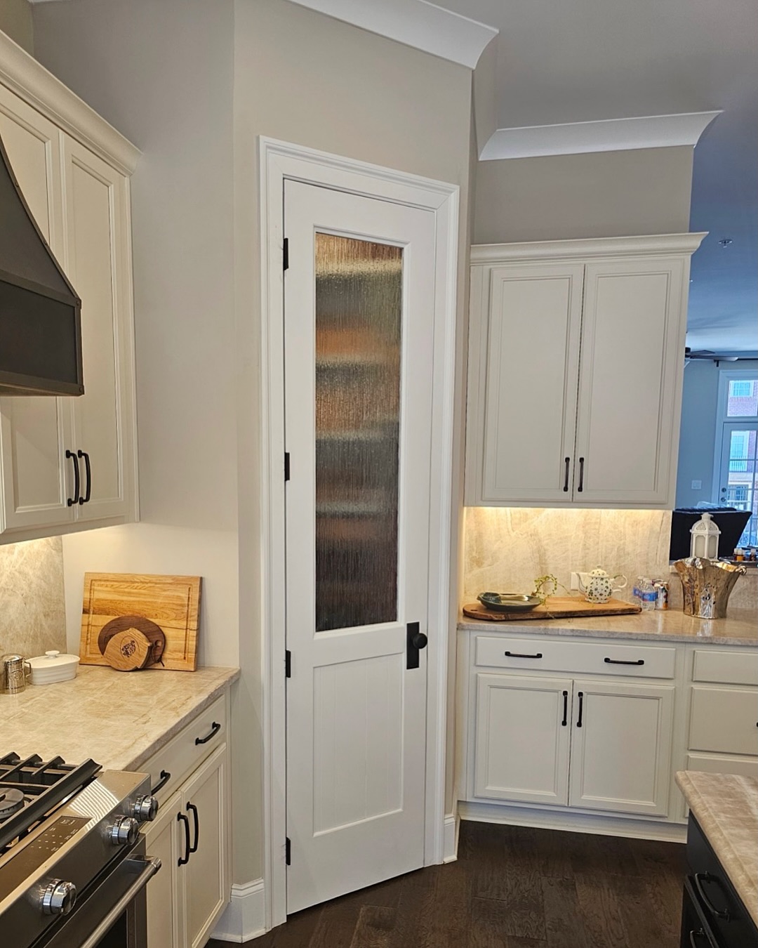 A modern kitchen design with white cabinets, a stove on the left, and a stylish pantry door with frosted glass in the center. Under-cabinet lighting illuminates the countertops, making it a great inspiration for pantry door ideas.