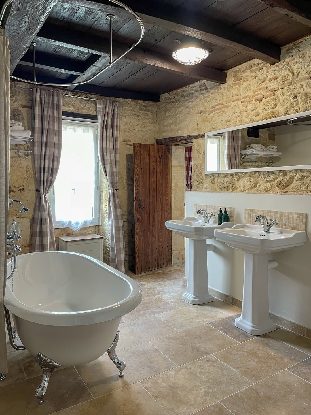 A rustic bathroom with a freestanding bathtub offers plenty of bathroom tub ideas, featuring twin pedestal sinks, stone walls, a tiled floor, and a window with plaid curtains for added charm.