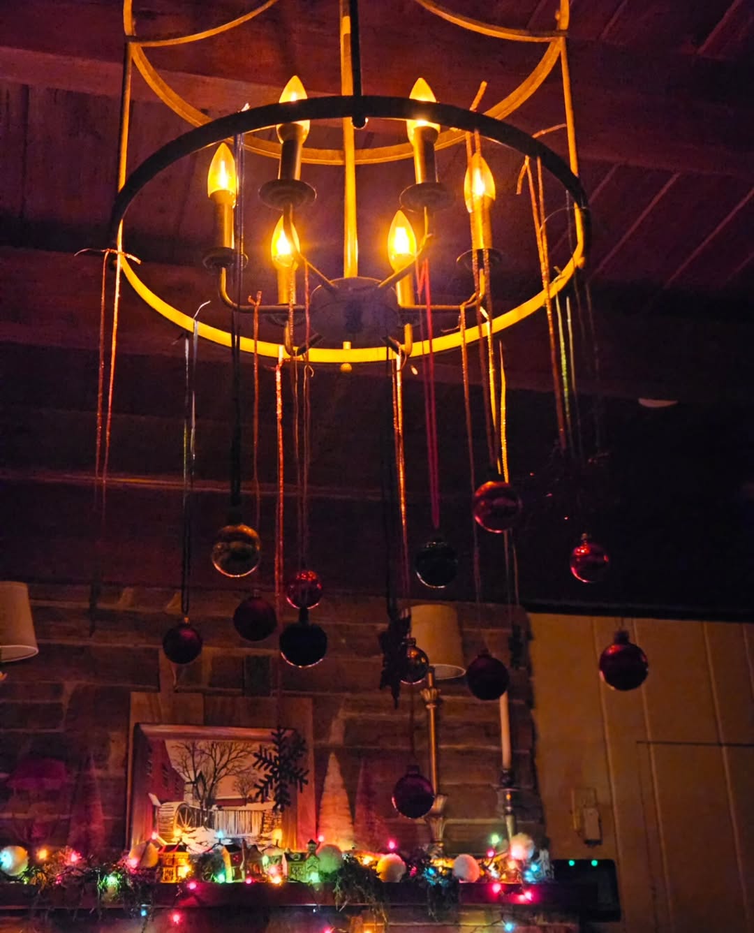 A chandelier with lit candle-shaped bulbs and colorful ornaments displays charming Christmas chandelier decor; festive lights and holiday chandelier decorations brighten the mantel below.