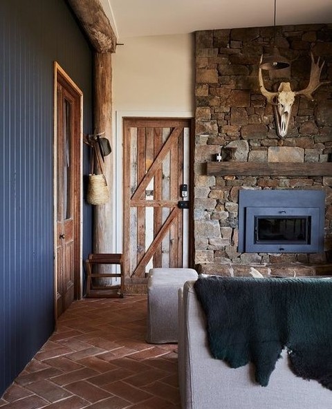 A rustic living room with ranch decor features a stone fireplace, wood doors, antler skull on the wall, herringbone brick floor, and a gray sofa draped with a dark green blanket.