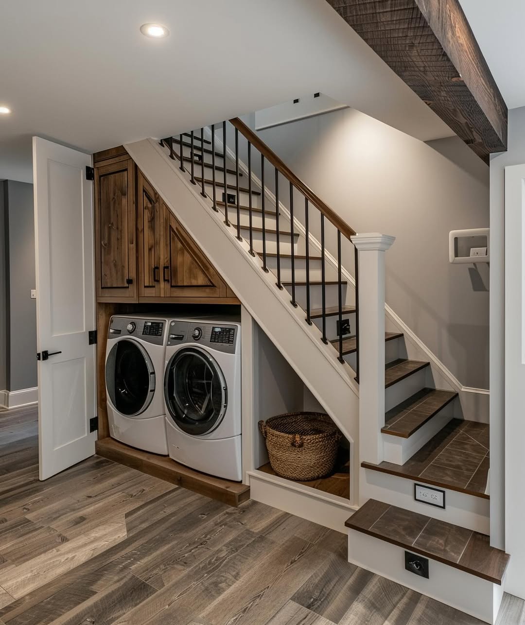 A stylish under stair laundry room features a washer and dryer tucked beneath the staircase, complemented by wooden cabinets, a laundry basket on the floor, wood flooring, and neutral wall colors.