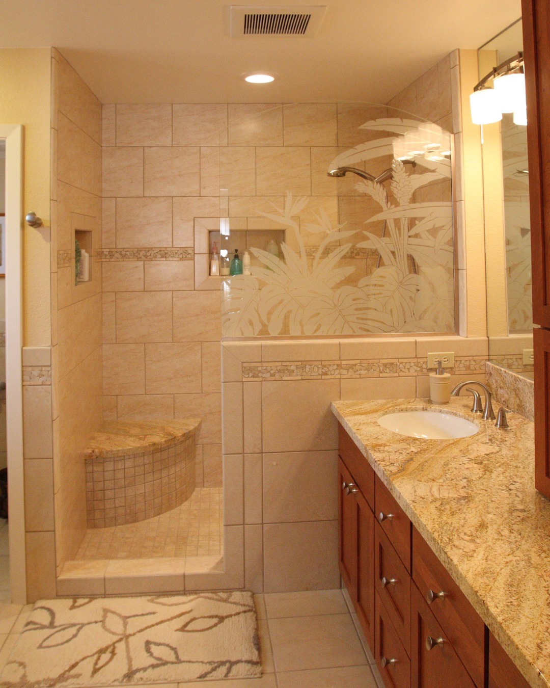 A bathroom with a no door shower featuring etched glass art, tiled bench, granite countertop, wooden cabinets, and a patterned rug—perfect inspiration for small bathrooms and walk-in shower ideas.