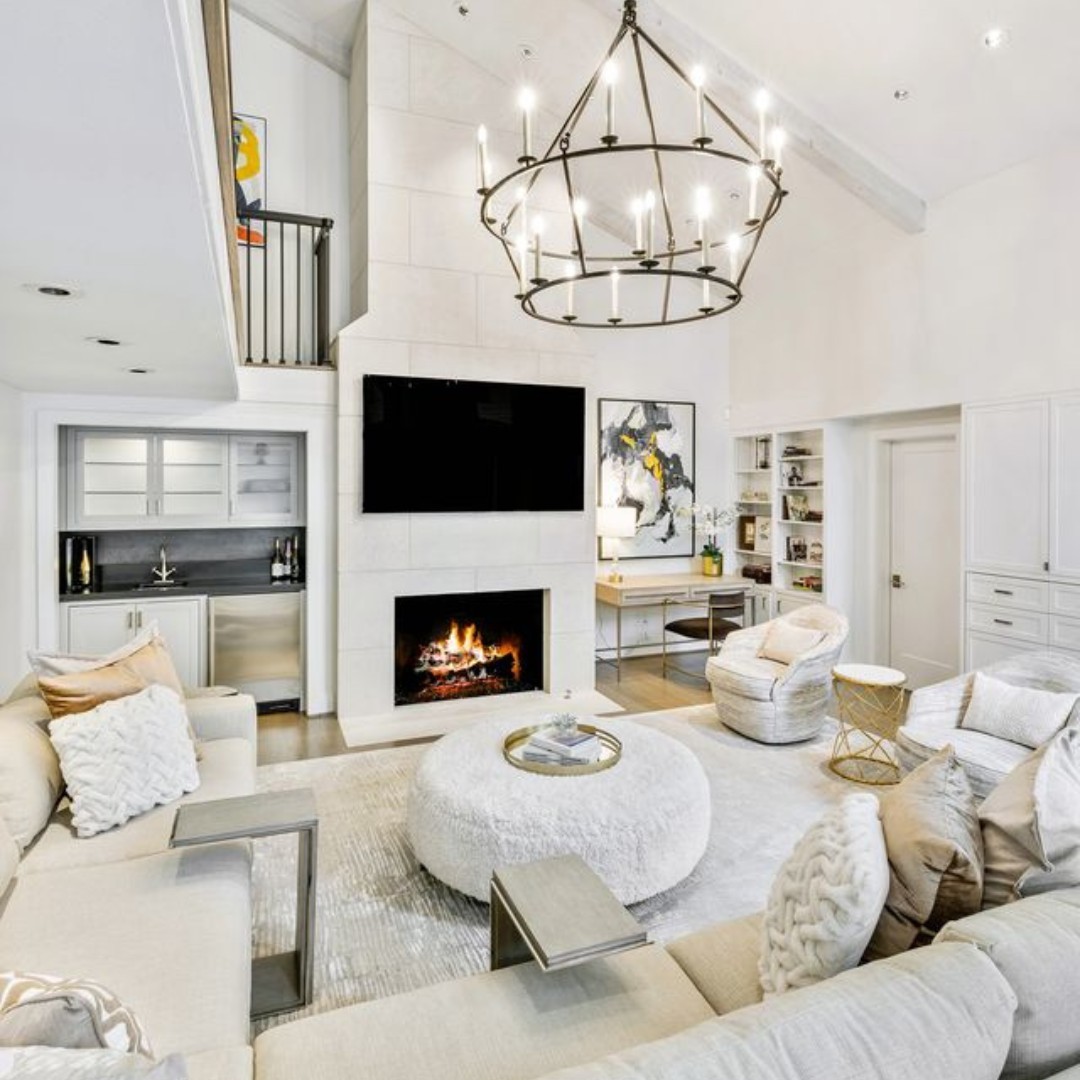 Modern high ceiling living room with neutral decor, sectional sofa, round ottoman, fireplace, large wall-mounted TV, built-in shelves, chandelier, and artwork—perfect for those seeking bigger home inspiration.