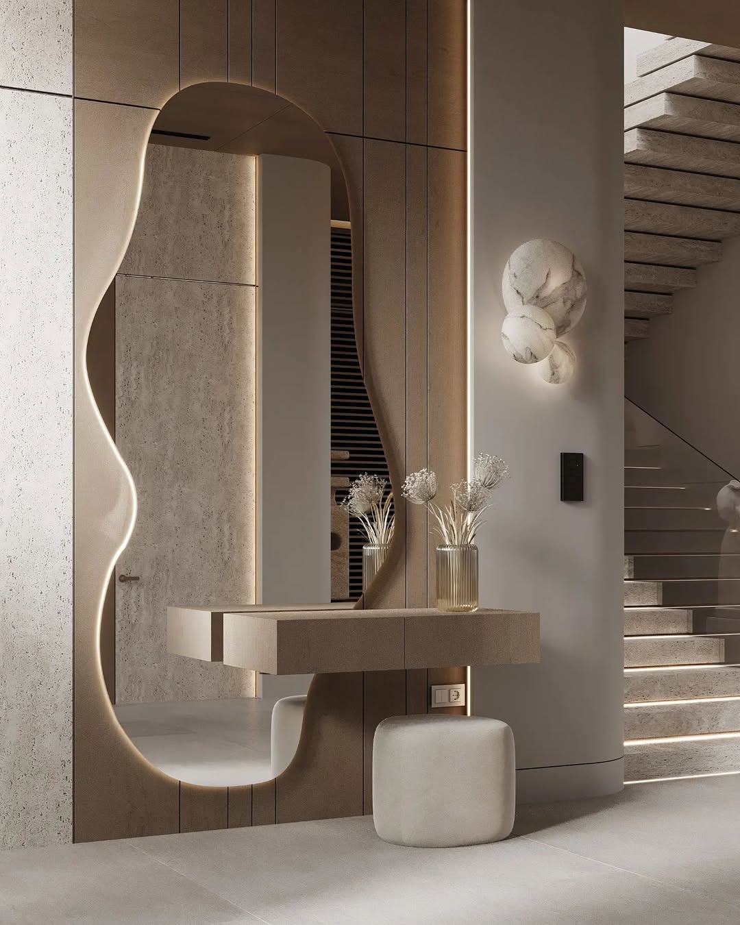 A modern foyer featuring a large irregular-shaped mirror, floating beige console with vases, a cube-shaped stool, and a staircase with minimalistic lighting—perfect for entryway decor inspiration.