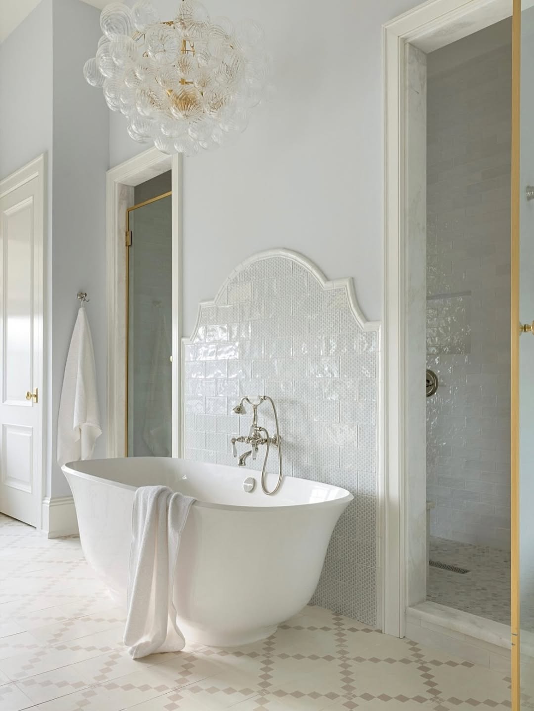 A freestanding white bathtub with a towel draped over the edge sits in a bright bathroom with white tile, glass shower, and a modern light fixture—perfect inspiration for fresh bathtub designs in your next bathroom remodel.