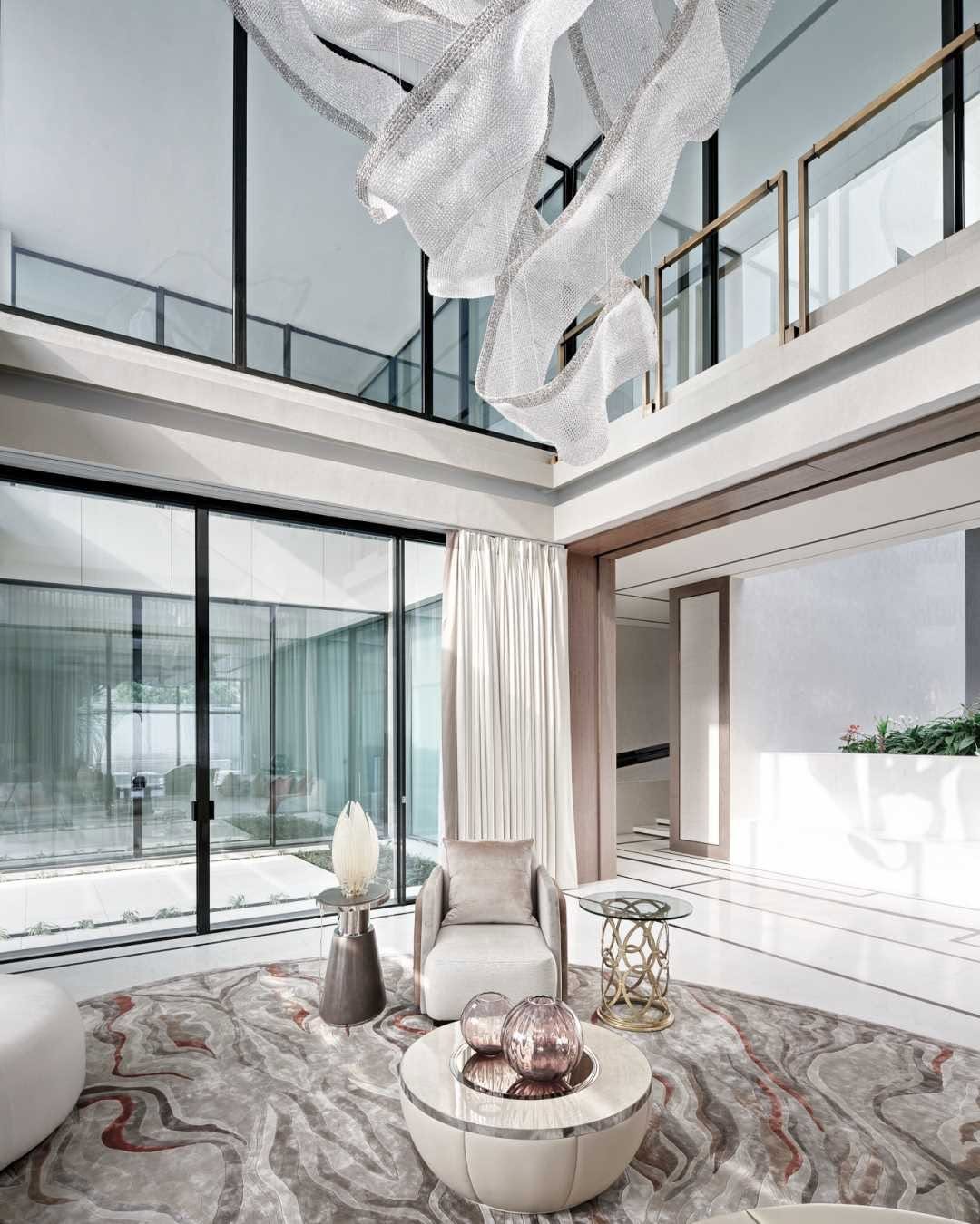 Modern living room with floor-to-ceiling windows, abstract chandelier, neutral furniture, and a patterned rug; natural light fills the airy, double-height space—perfect for high ceiling living room ideas.