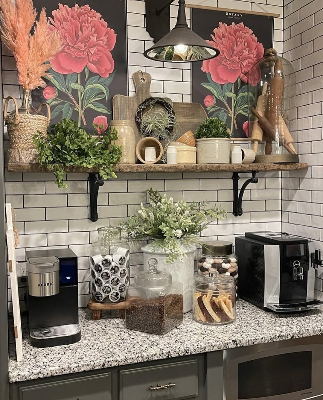 A coffee bar featuring two coffee machines, jars of beans and pastries, mugs, plants, and decorative floral art on a white tile backsplash—perfect Kitchen Coffee Bar Ideas to bring order to your Countertop Chaos.