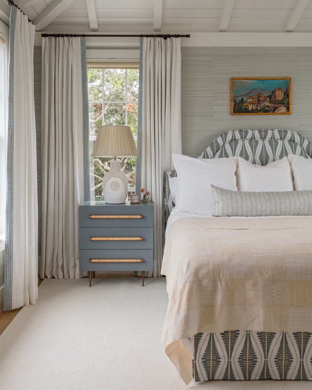 Bedroom with a patterned bed, light beige bedding, blue nightstand with lamp, large window with curtains, and a landscape painting on the wall offers fresh Bedroom Interior Ideas for a cozy and stylish retreat.