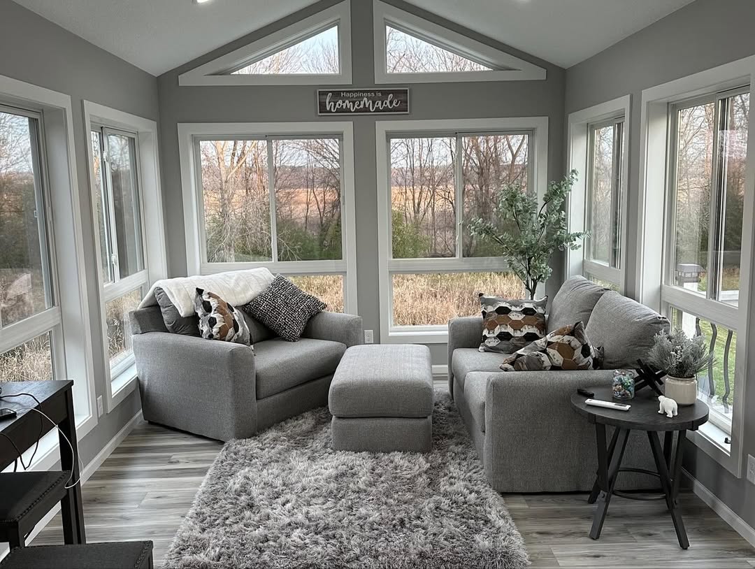A sunroom with large windows, a gray armchair and sofa, a matching ottoman, side table, and plush rug on light wood flooring—perfect for inspiring 3 season porch ideas. Pillows and a throw blanket add cozy accents to the inviting decor.