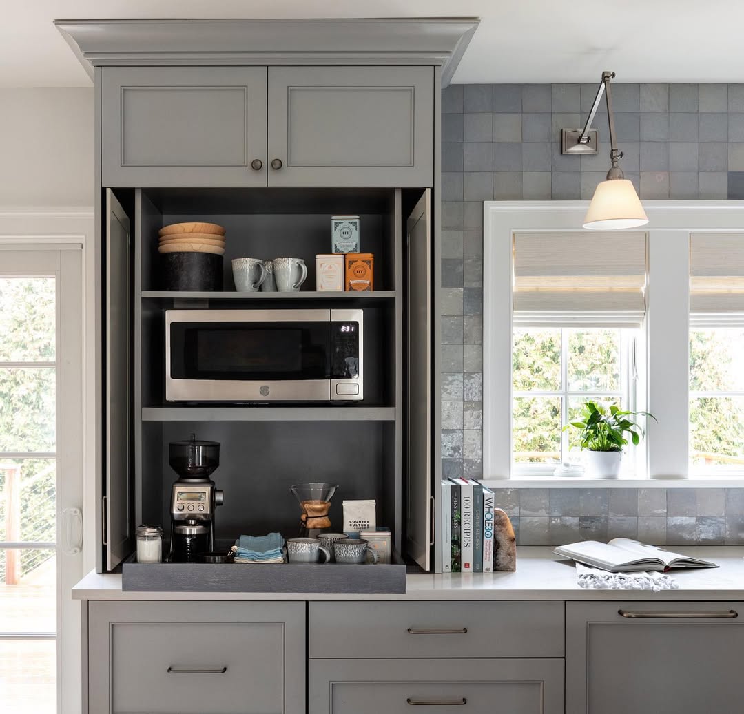 A modern built-in kitchen coffee bar with a microwave, coffee maker, cups, coffee supplies, books, and a window with natural light—perfect inspiration for those seeking sleek kitchen coffee bar ideas without countertop chaos.