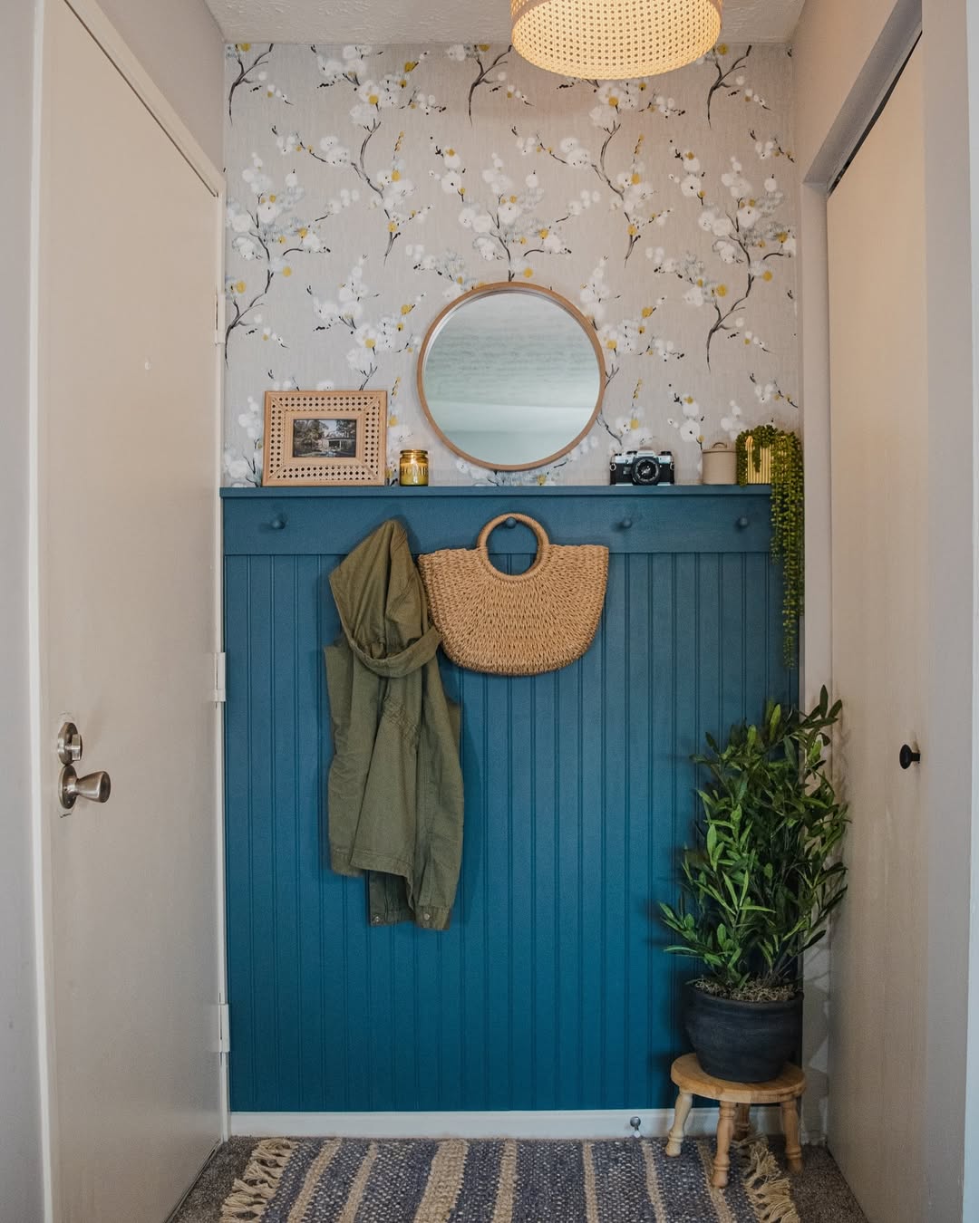 Charming small entryway with blue wainscoting, wall hooks for a jacket and woven bag, round mirror, wallpaper accent, potted plant, and framed photo on a shelf.