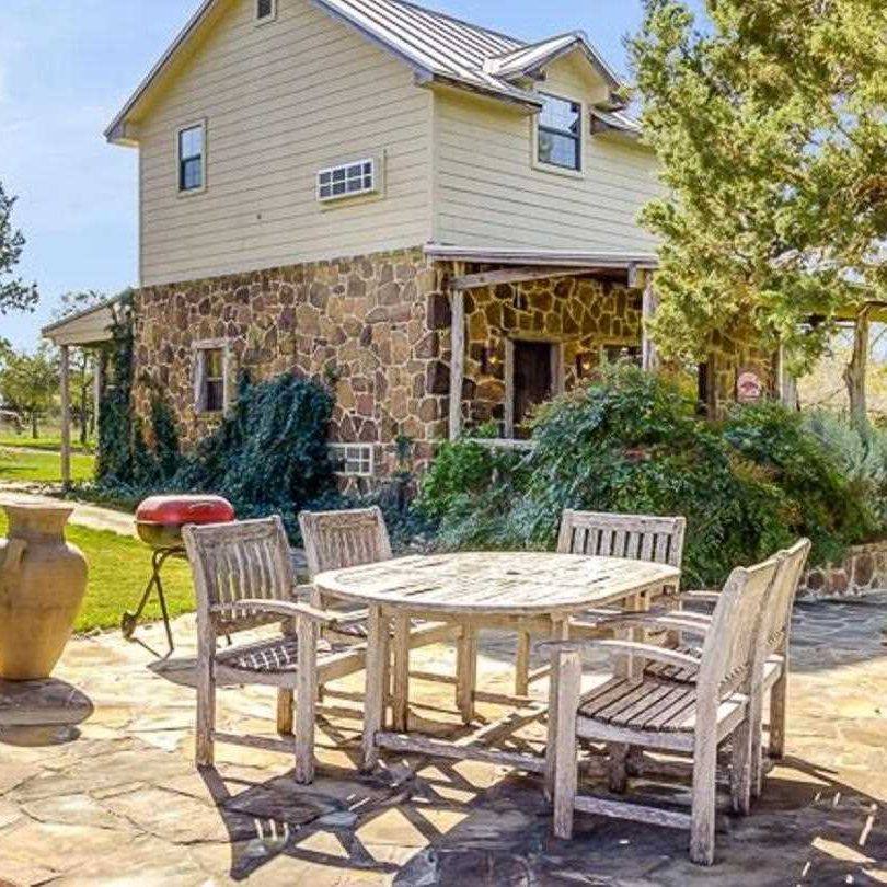 A wooden outdoor table with four chairs sits on a stone patio in front of a stone and wood house, showcasing ranch decor with a covered porch and lush surrounding greenery.