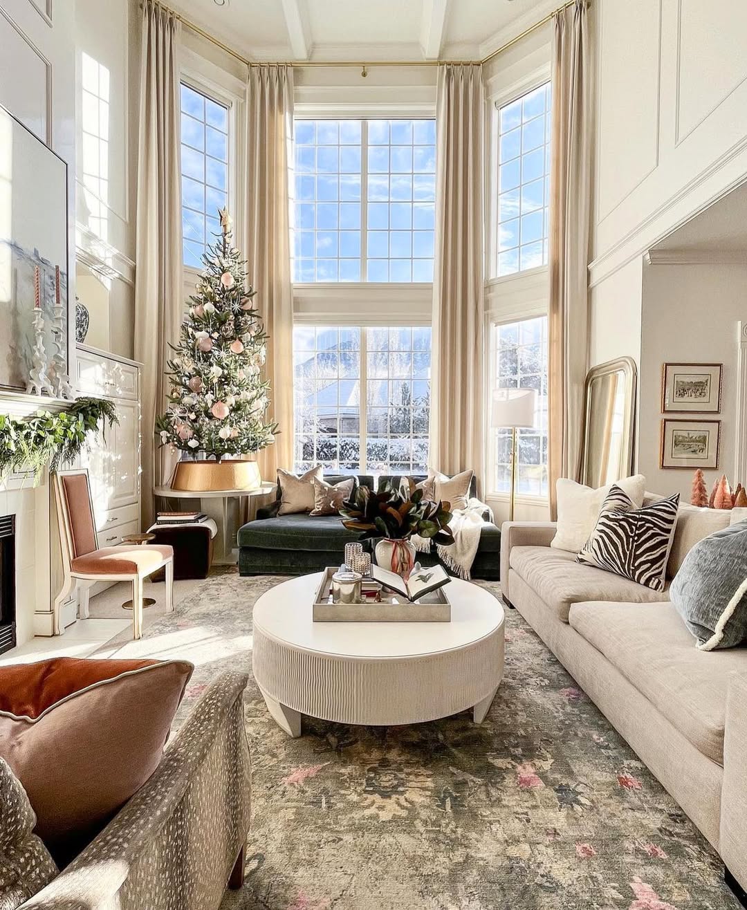 A bright, high ceiling living room with large windows, a Christmas tree, beige and gray sofas, a round coffee table, and neutral-toned decor on a patterned rug. Snow is visible outside—perfect for those seeking bigger home inspiration.