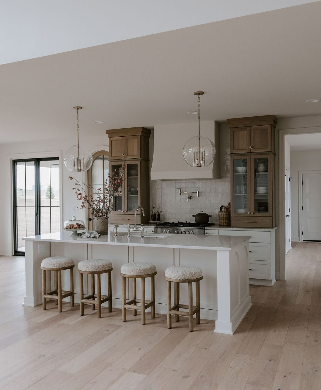 Modern kitchen with light wood floors, a white island with four cushioned stools, pendant lights, and cabinetry with glass-front doors. Perfect for those seeking open kitchen ideas, branches in a vase add a touch of nature to the countertop.