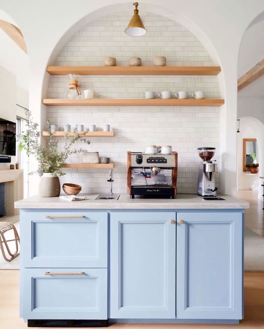 A modern kitchen coffee bar with light blue cabinets, open wooden shelves displaying mugs and bowls, a coffee machine, and a white tile backsplash—perfect inspiration for those seeking stylish kitchen coffee bar ideas.