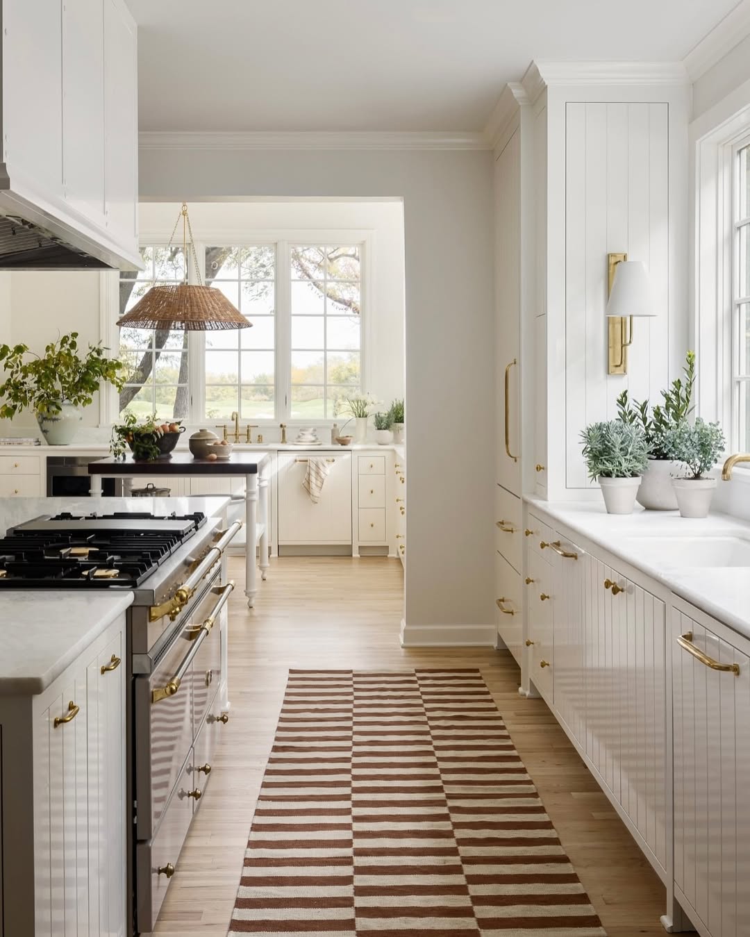 A modern galley kitchen with white cabinets, gold hardware, a striped rug, potted plants, and large windows flooding the space with natural light—perfect for small kitchen ideas.