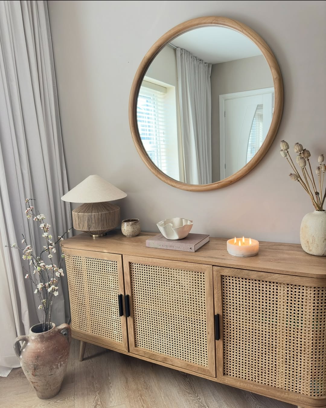 A wooden sideboard with rattan doors sits against a wall, topped with sideboard decor and a lit candle. A round mirror hangs above, reflecting a window with white curtains—perfect inspiration for sideboard decorating.