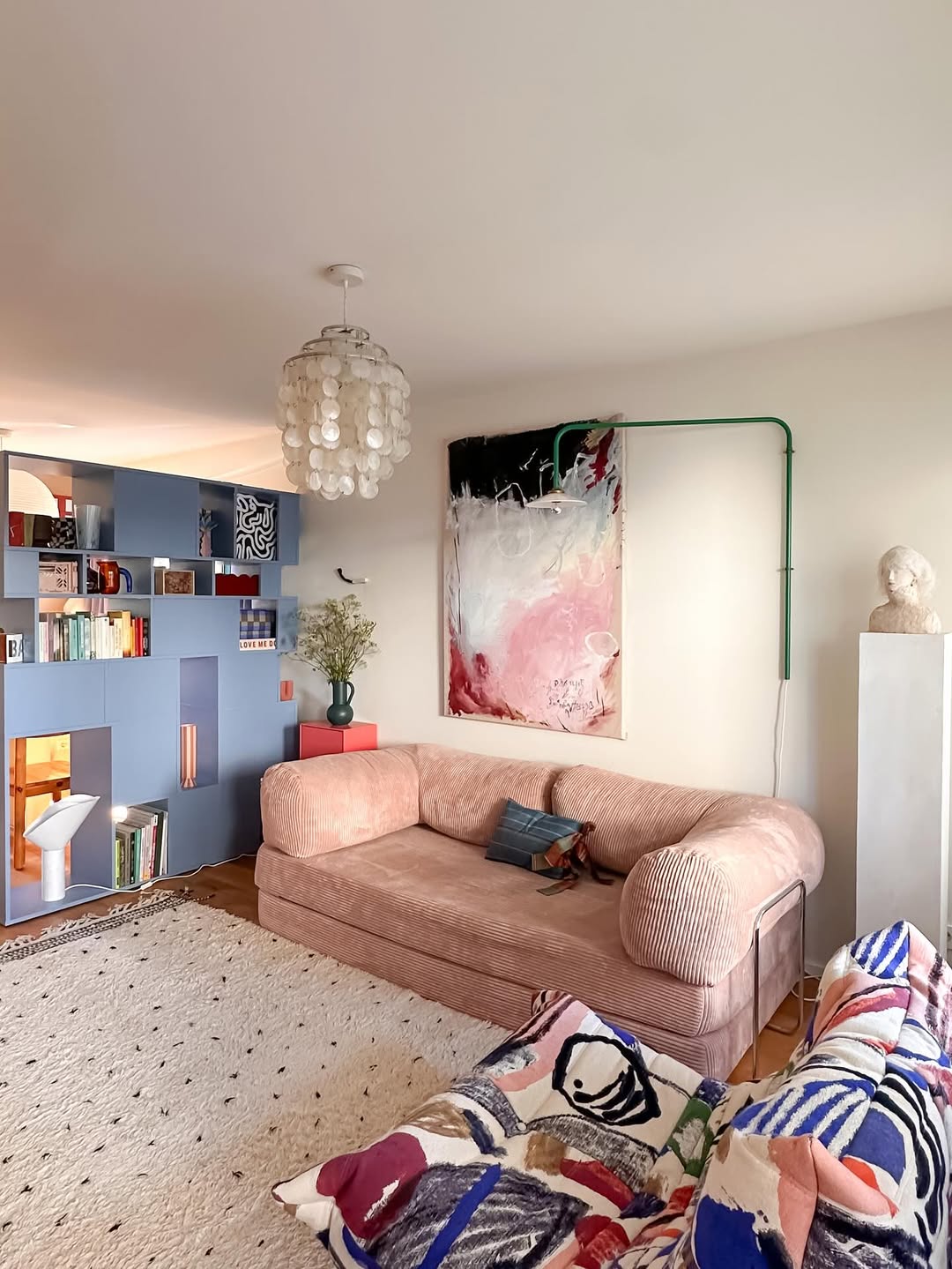 A modern living room with a pink velvet sofa, abstract wall art, a light fixture, and a blue geometric bookshelf showcases tiny apartment ideas, featuring space-saving tips like colorful pillows and a patterned rug for a cozy, stylish look.
