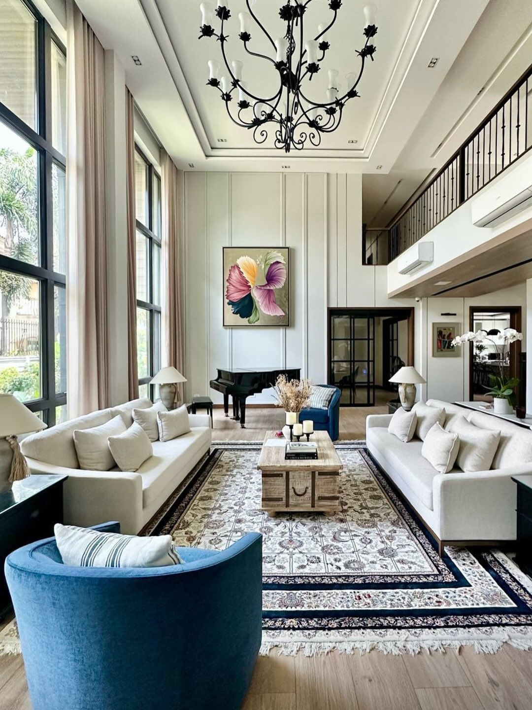 Spacious high ceiling living room with tall windows, two beige sofas, two blue chairs, a grand piano, a large chandelier, and a colorful abstract painting—a perfect inspiration for bigger home design.