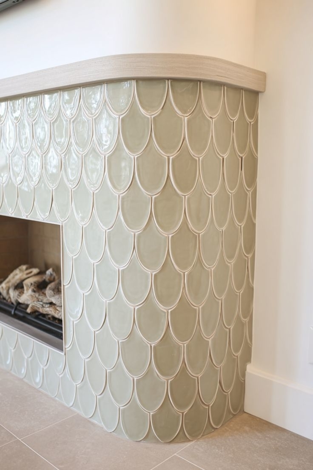 A fireplace with a curved exterior covered in glossy, light green fish scale tiles and a light wood mantel offers fresh fireplace tile ideas for a modern, stylish space.