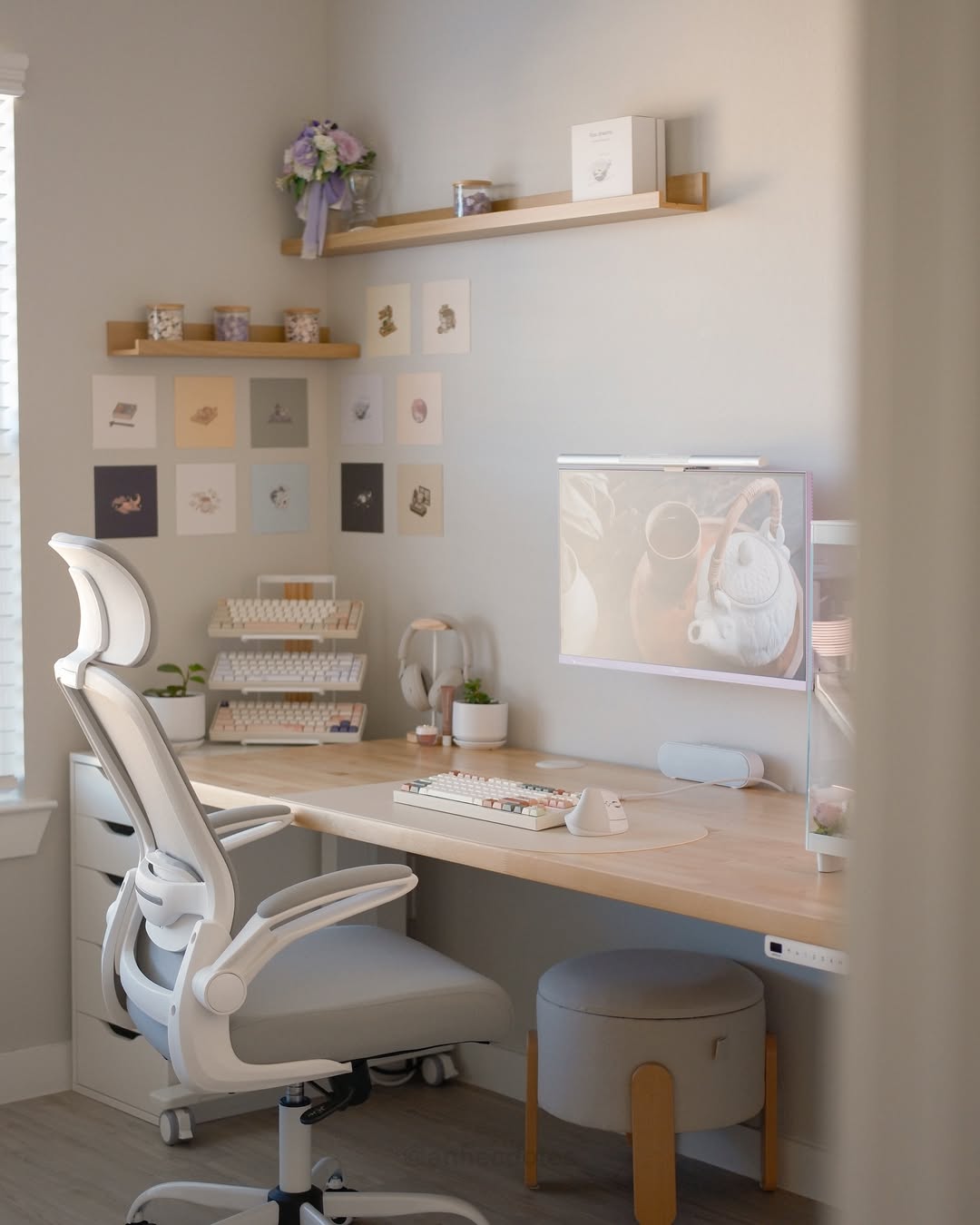 A modern home office with a large monitor, white ergonomic chair, wooden desk, wall art, and shelves showcasing desk organization ideas and storage items—all in neutral and pastel tones.