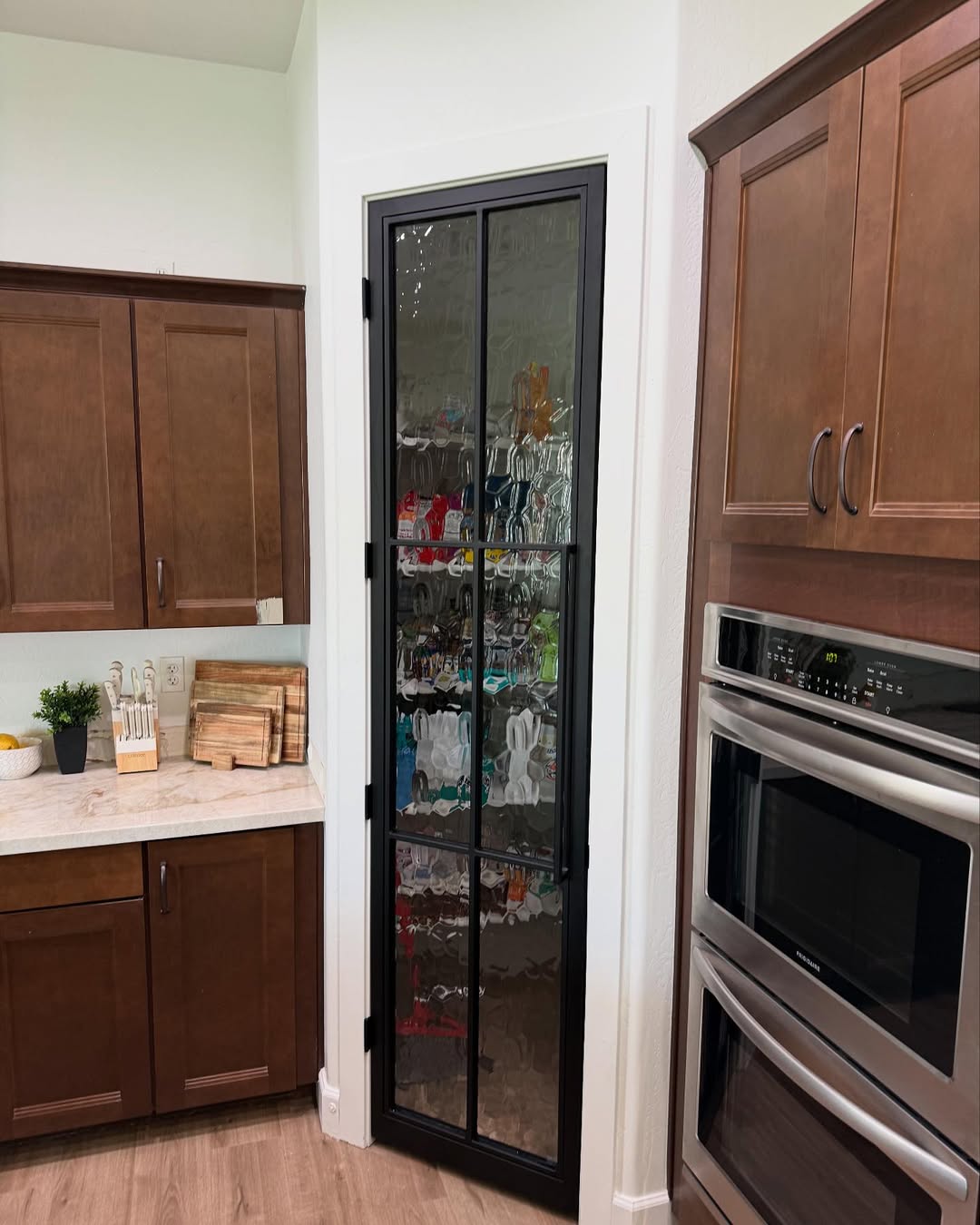A kitchen pantry with stylish pantry doors and a glass panel displays various food items inside; brown cabinets, a double oven, and a countertop are visible nearby.